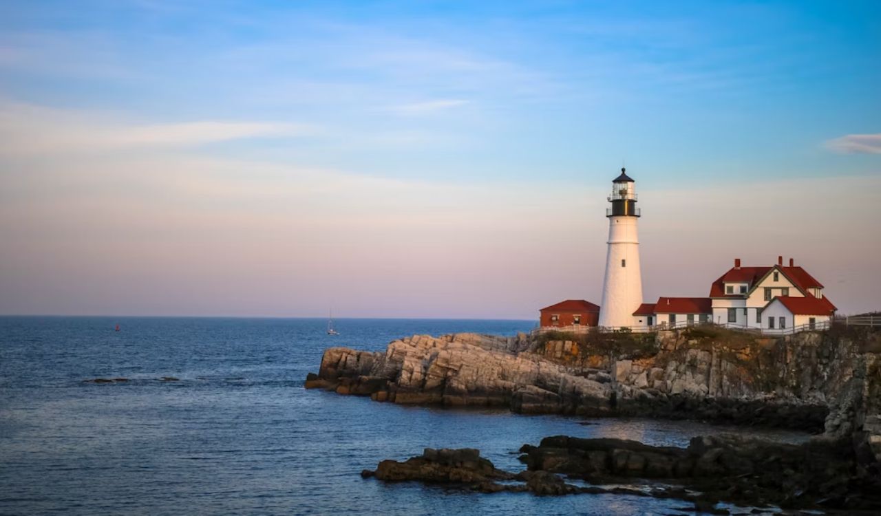 A white lighthouse with an attached building stands on a rocky coast beneath a blue and pink sunset sky.