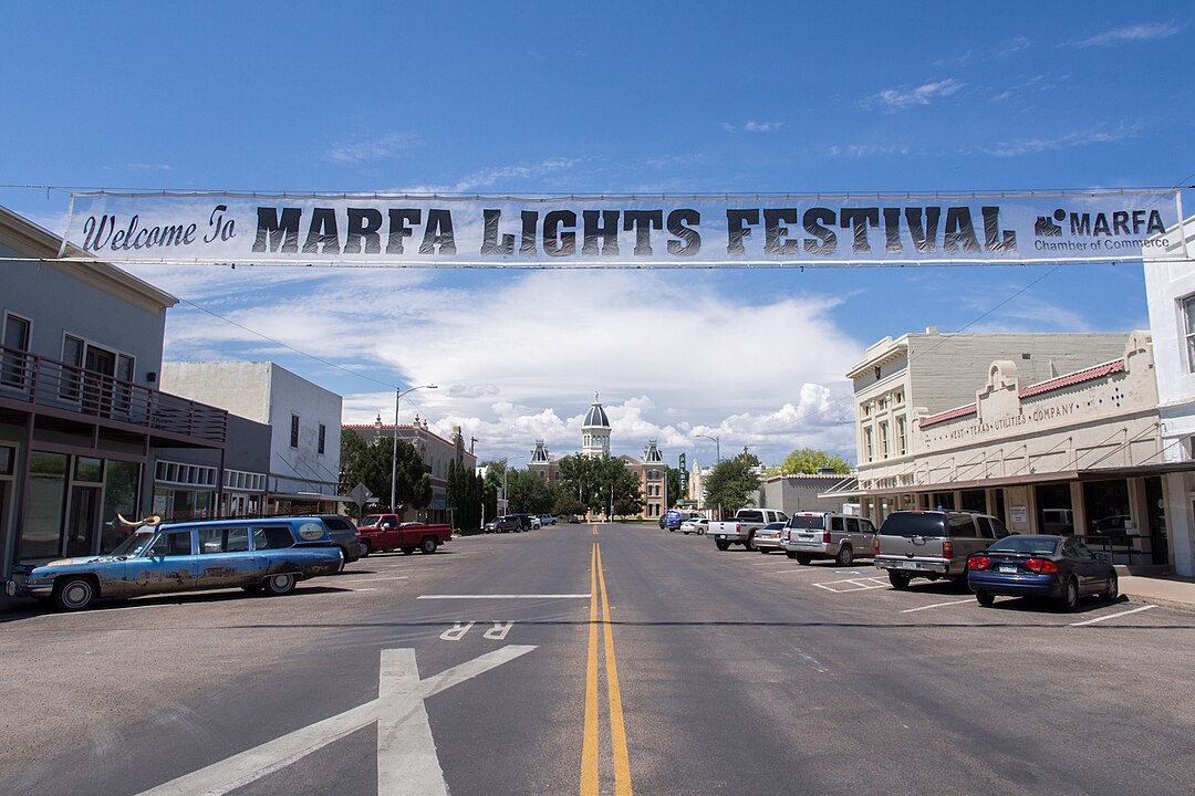 A street in Marfa, Texas displays a "Welcome to Marfa Lights Festival" banner overhead with cars parked on both sides.