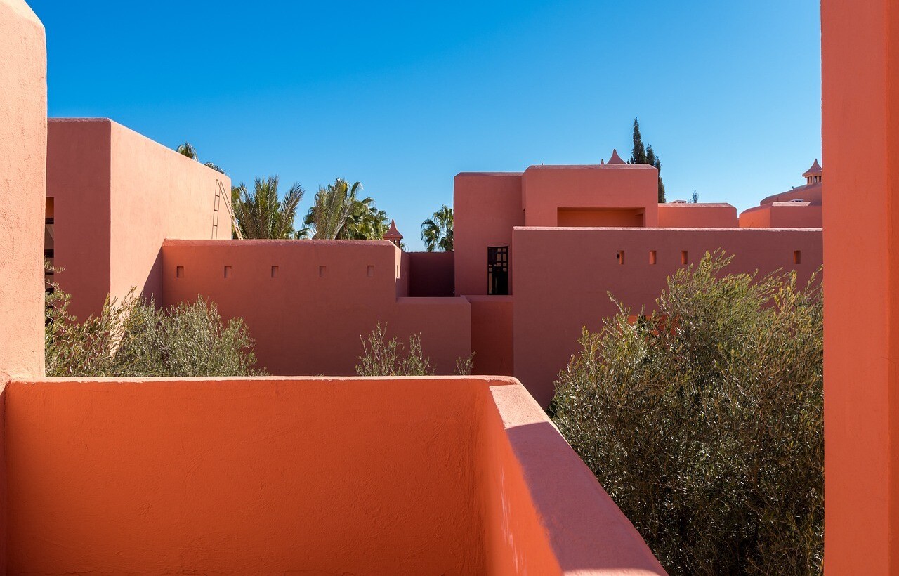 Coral buildings with flat roofs and few windows stand beneath a clear blue sky, trees in the foreground.