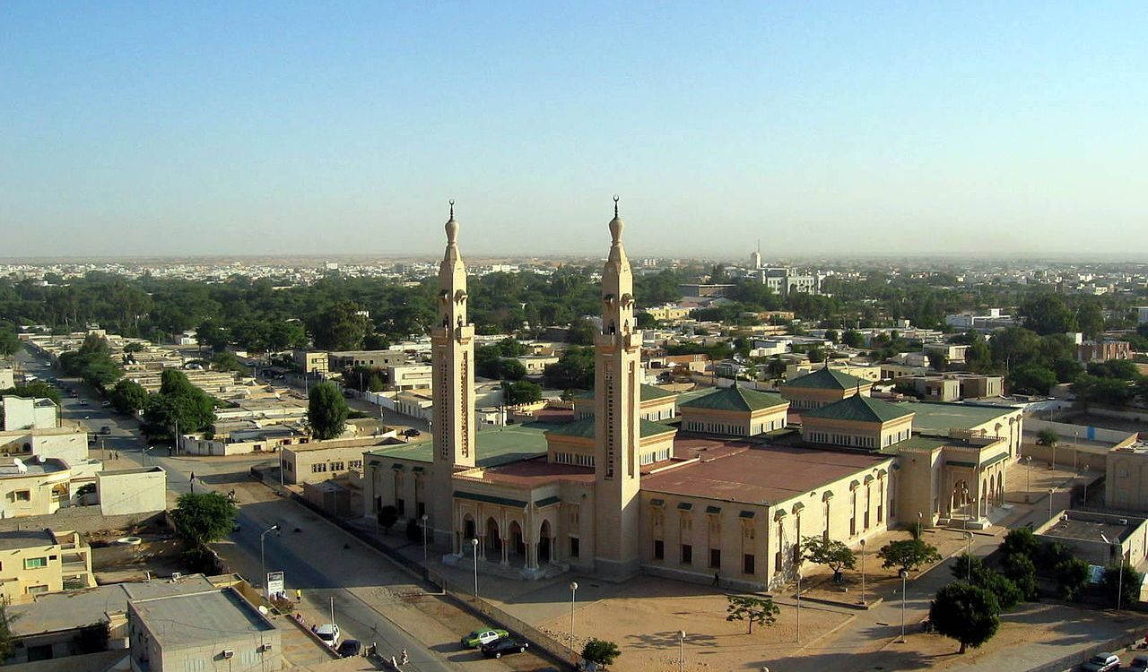 A large mosque with two tall minarets stands on a city corner among low-rise buildings and greenery.