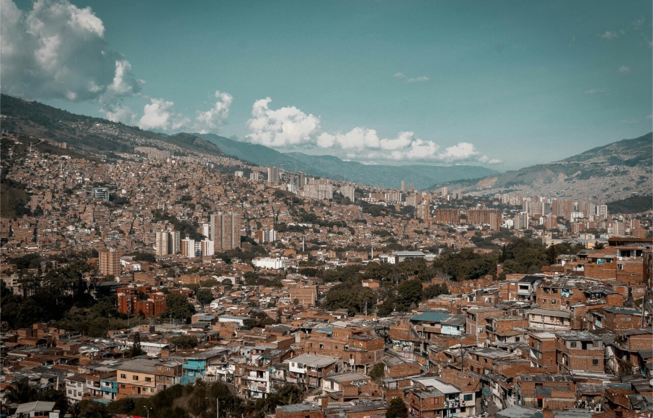A dense urban area with mixed low- and high-rise buildings lies beneath a partly cloudy sky, surrounded by mountains.