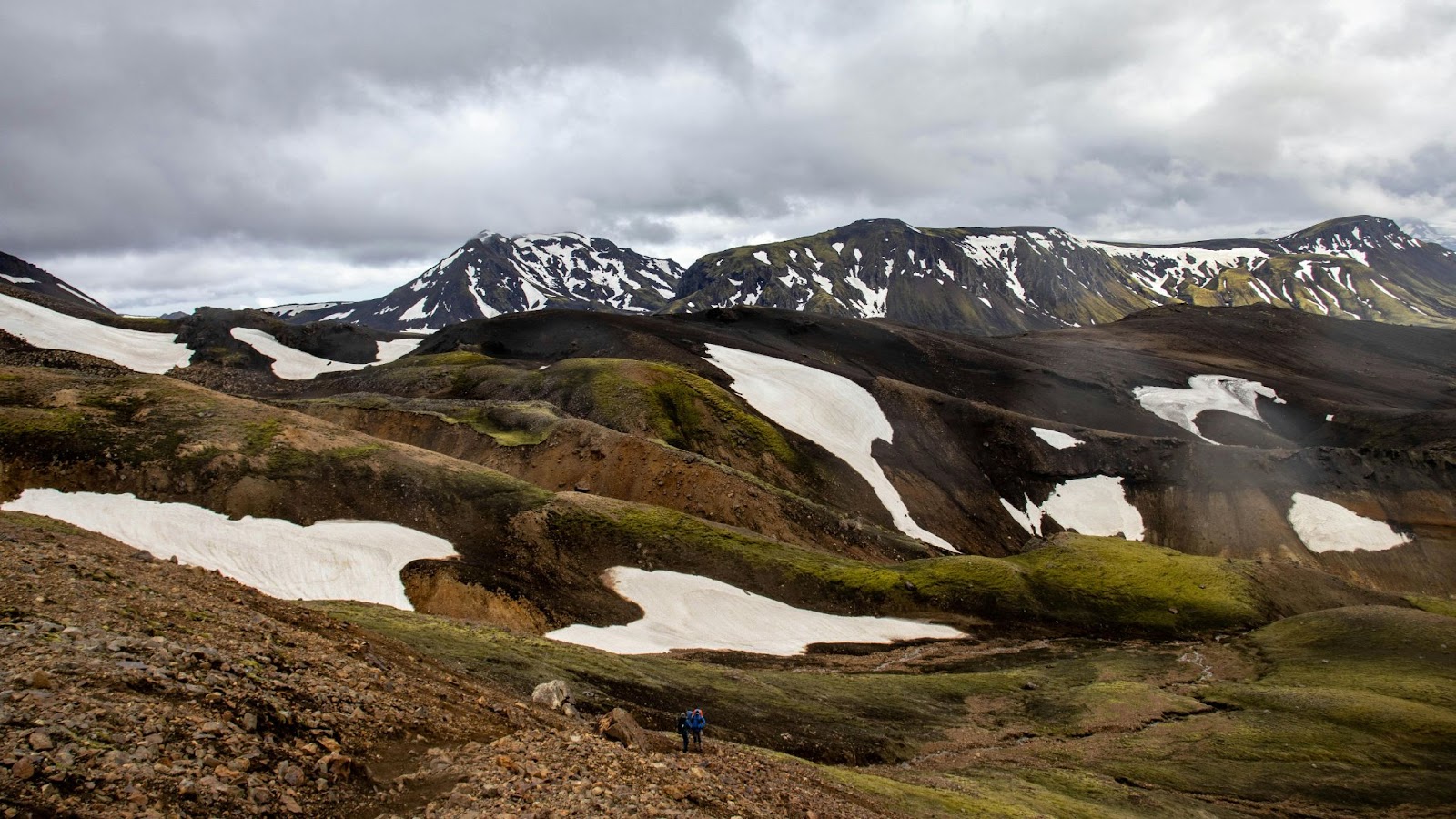 Hikers trek beneath a cloudy sky across a rugged volcanic landscape dotted with snow patches and backed by mountains.
