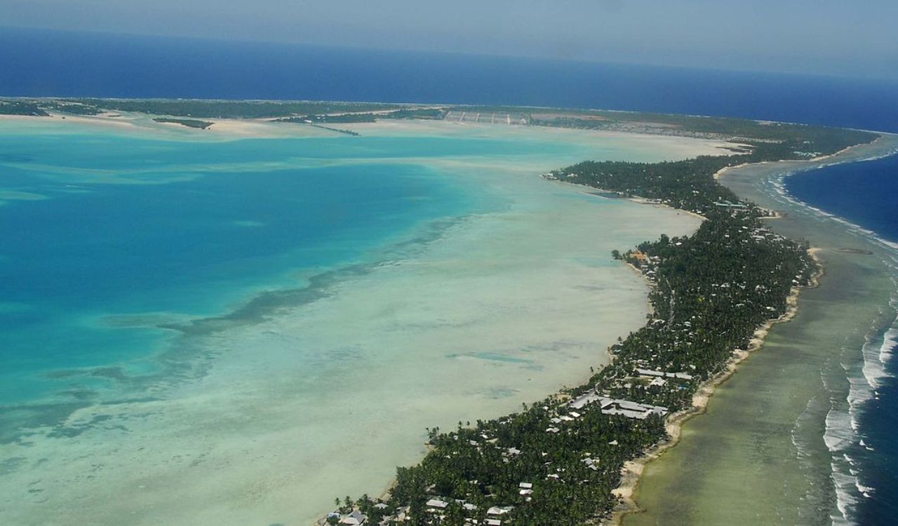 Aerial view of an atoll with a turquoise lagoon, white sandy shores, scattered buildings, and surrounding ocean.