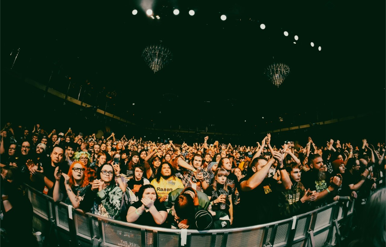 A large crowd stands behind a barricade at an indoor concert, many raising their hands or clapping under stage lights.