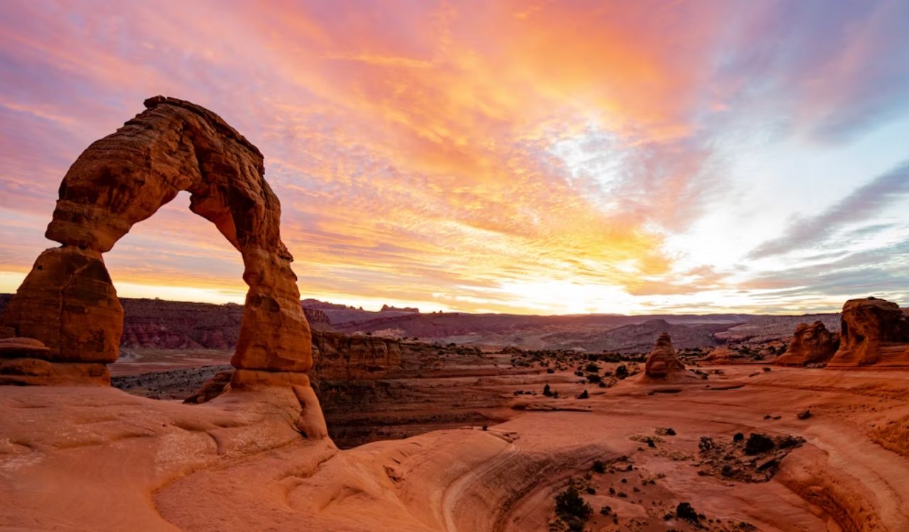 A sandstone arch rises from a rocky desert beneath a vibrant sunrise sky.