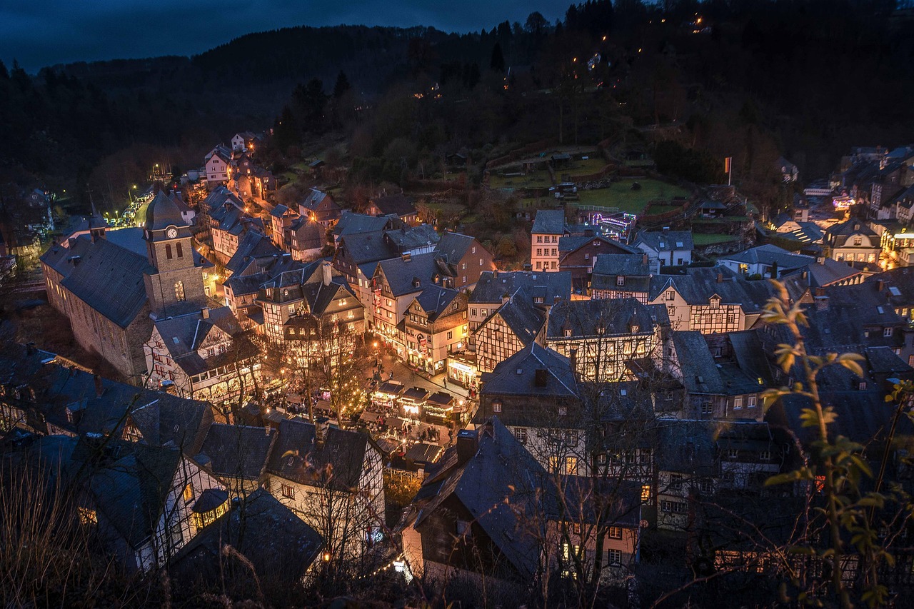 Aerial view of illuminated half-timbered houses in a small town at dusk, surrounded by hills and trees.