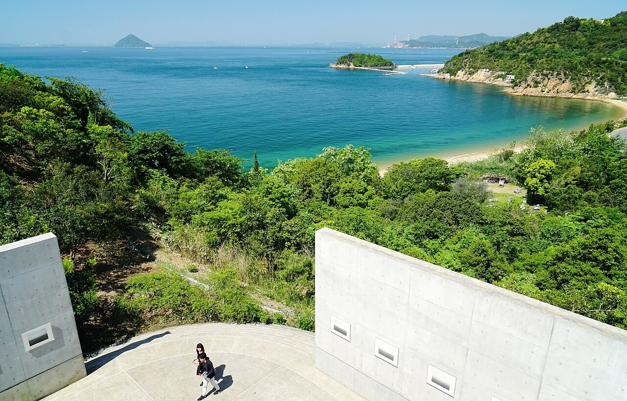 A person walks on a concrete path overlooking greenery, a sandy beach, and a calm blue sea with distant islands.