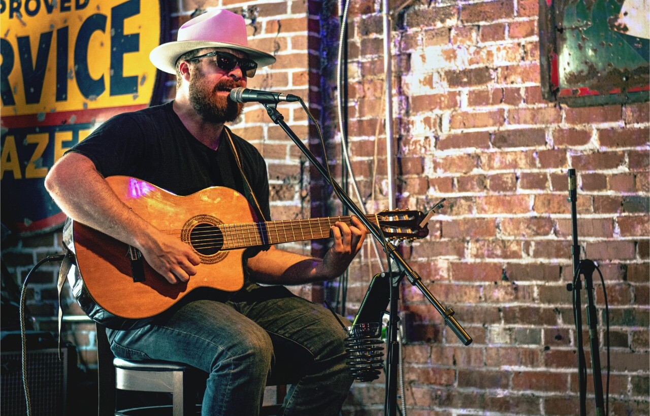 A man in sunglasses and a hat plays acoustic guitar and sings into a microphone before a brick wall indoors.