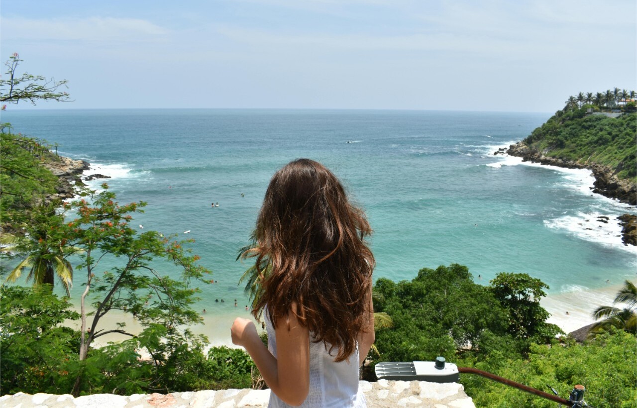 A woman with long brown hair stands on a stone terrace overlooking a tropical beach and turquoise ocean beneath a partly cloudy sky.