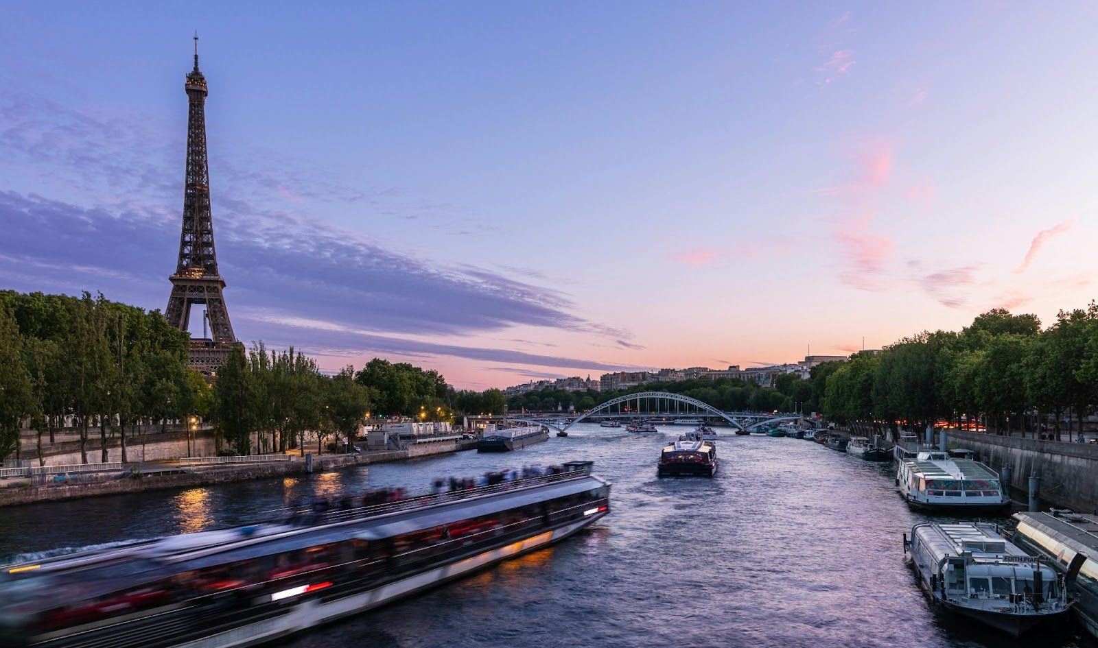 At dusk, the Eiffel Tower overlooks the Seine River, where boats glide past a distant bridge.