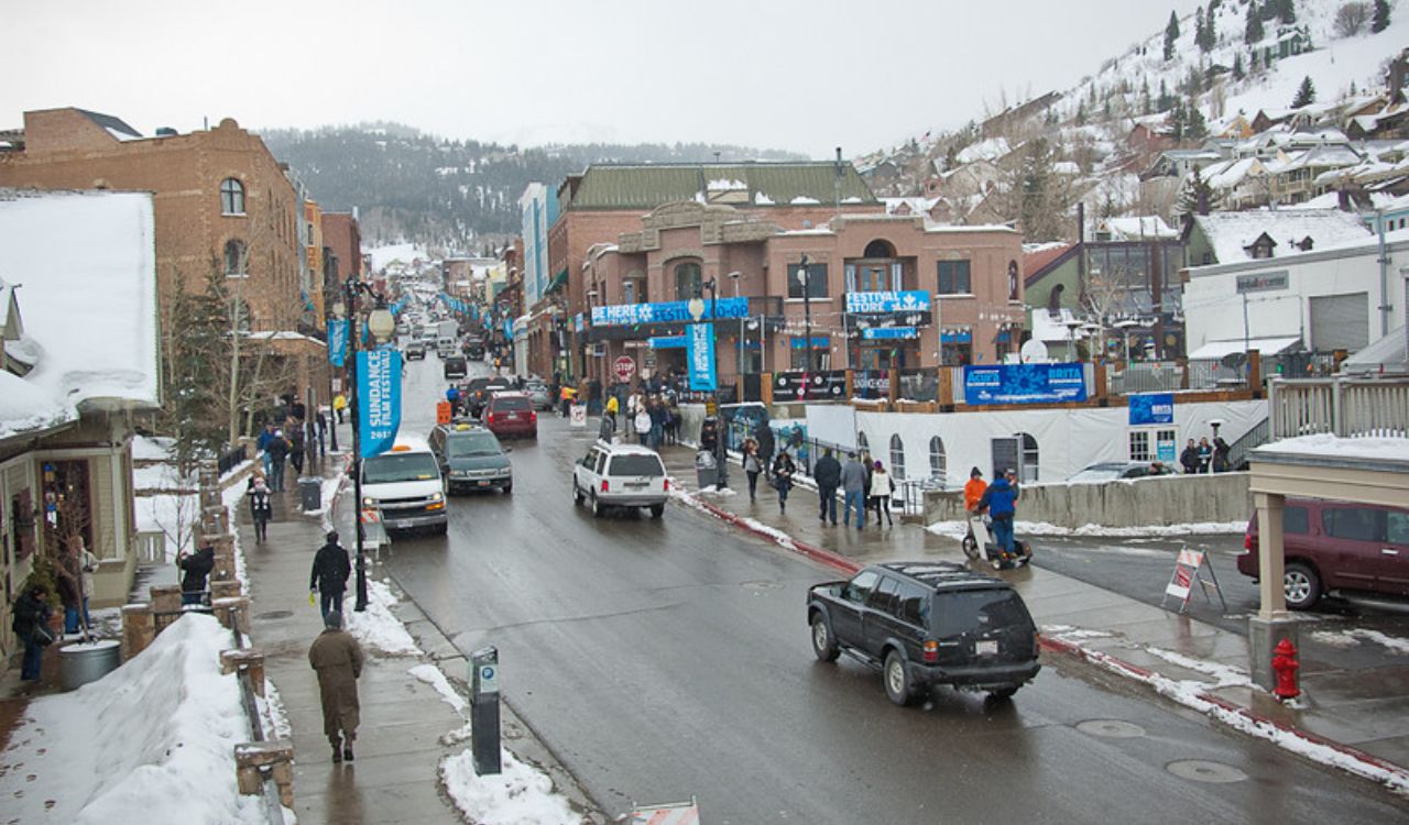 A snowy main street in a small mountain town features cars, pedestrians, banners, buildings, and snowy hills in the background.