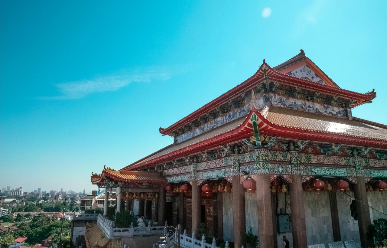 An ornate Chinese temple with red roofs overlooks a cityscape beneath a clear blue sky.