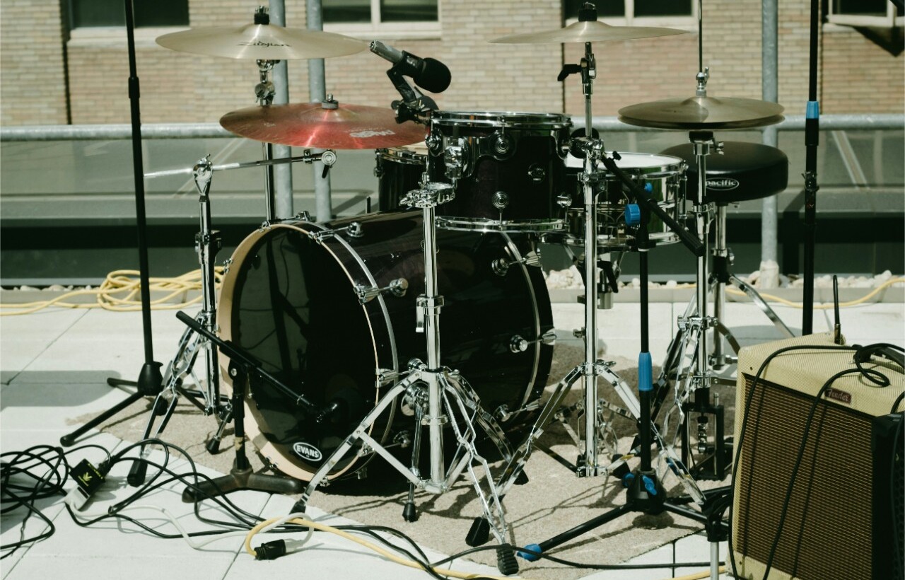 A black drum kit with cymbals and microphones is set up outdoors on a rooftop beside an amplifier and yellow cables.