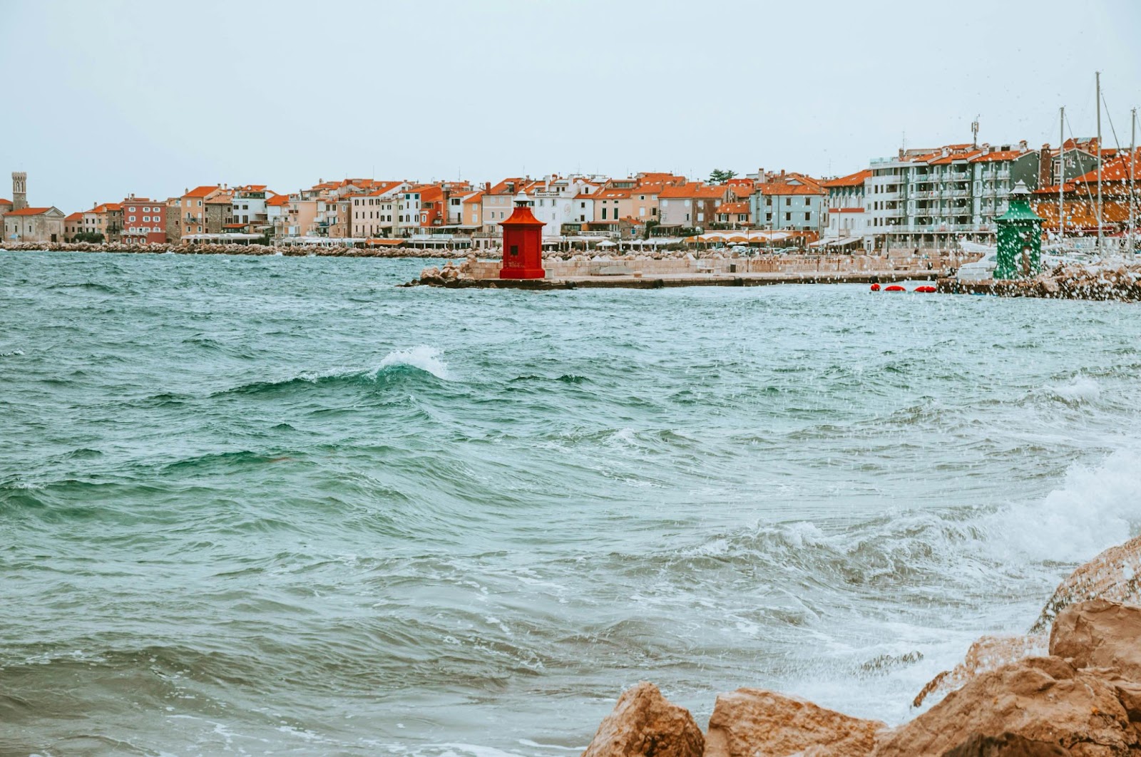 Waves crash against a rocky shore near a red lighthouse, with orange-roofed town buildings in the background.