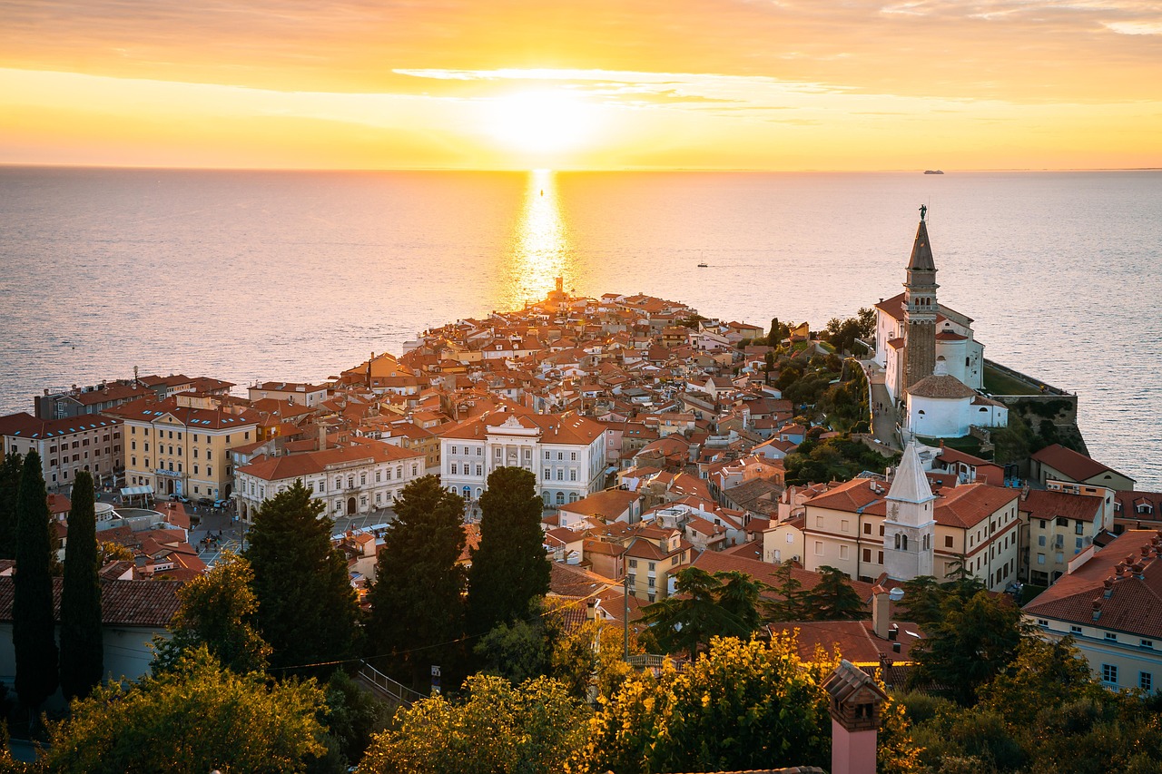 At sunset, sunlight reflects on the calm sea beside a coastal town with red-roofed buildings and a prominent church tower.