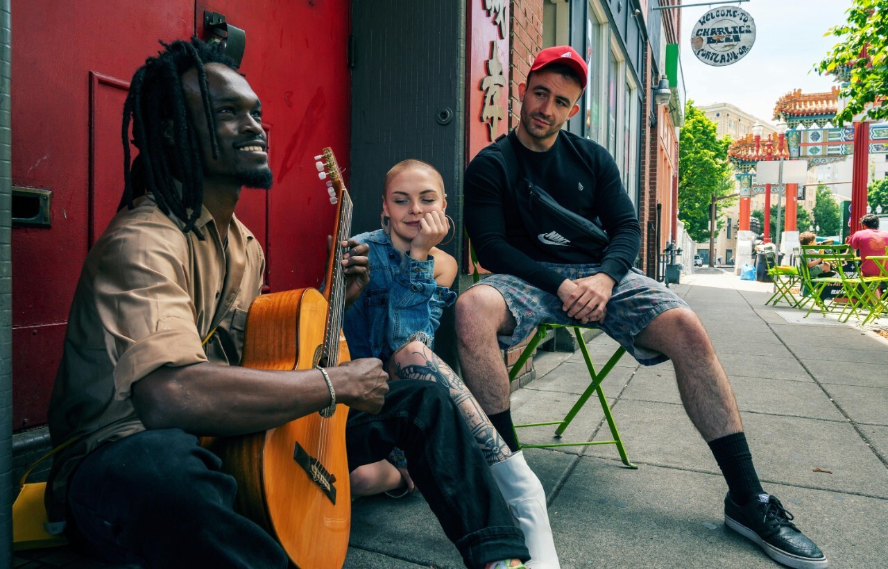 Three people sit on a city sidewalk among outdoor tables; one plays guitar while the others listen.
