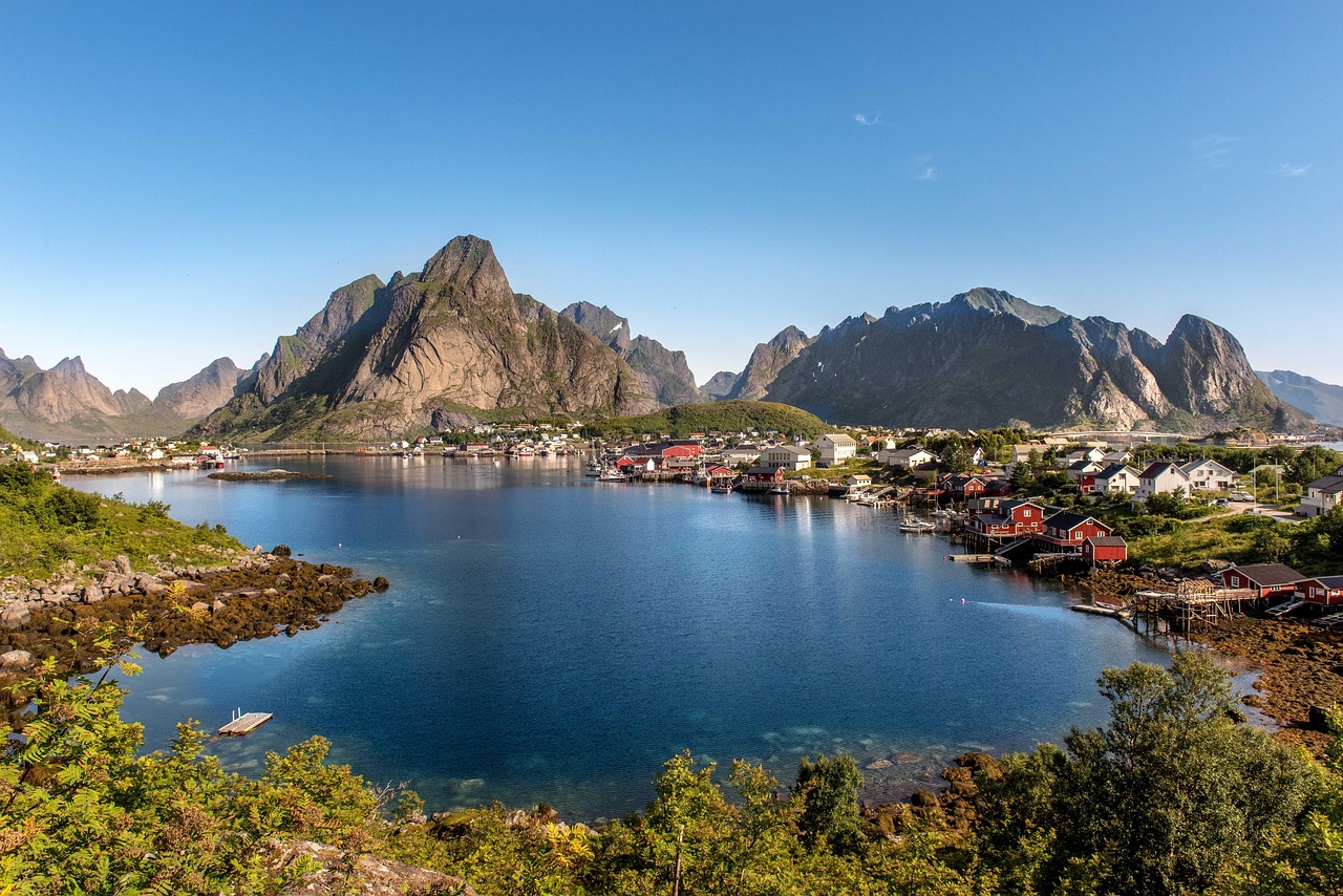 A calm fjord bordered by steep mountains features a waterfront village of red and white houses beneath a clear blue sky.