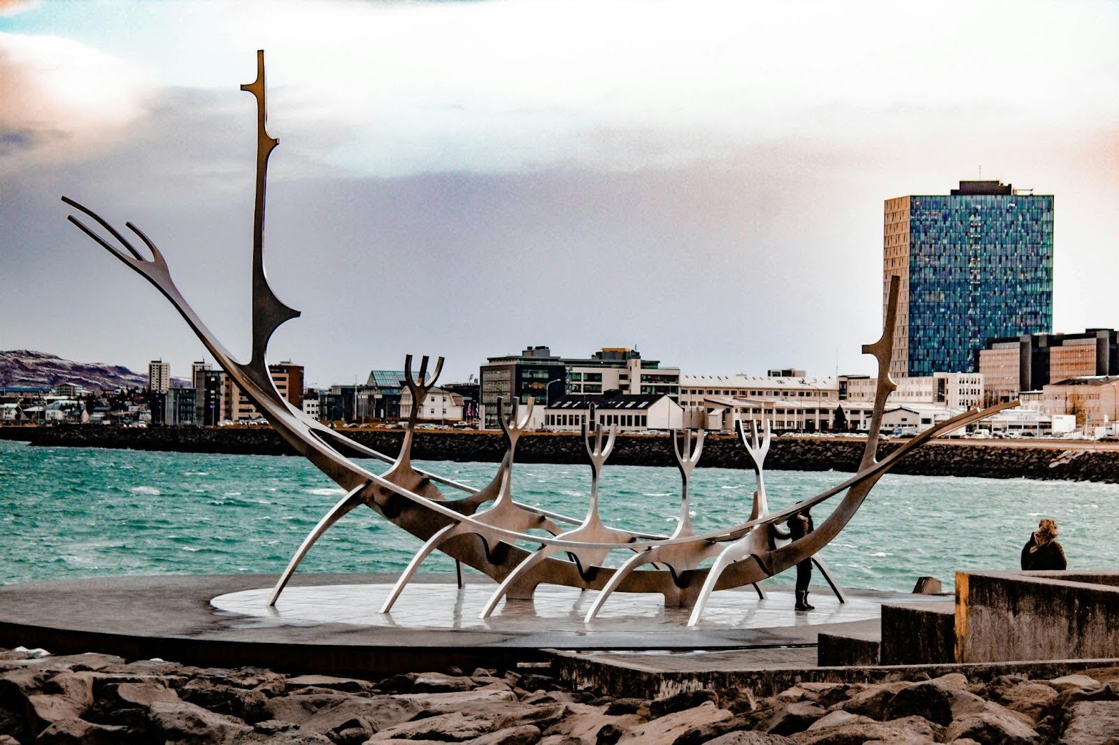 A large metallic Viking ship sculpture stands by the waterfront, backed by city buildings, mountains, and a cloudy sky.
