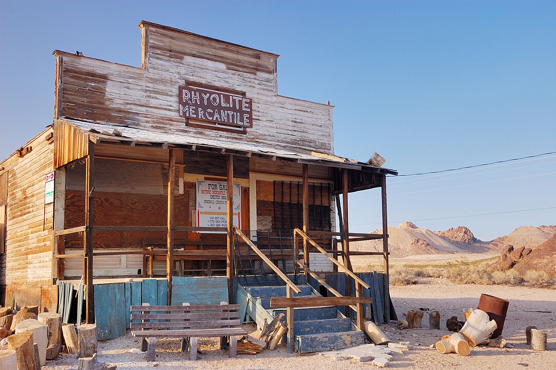 A weathered wooden building labeled "Rhyolite Mercantile" stands in a deserted arid landscape with mountains behind.