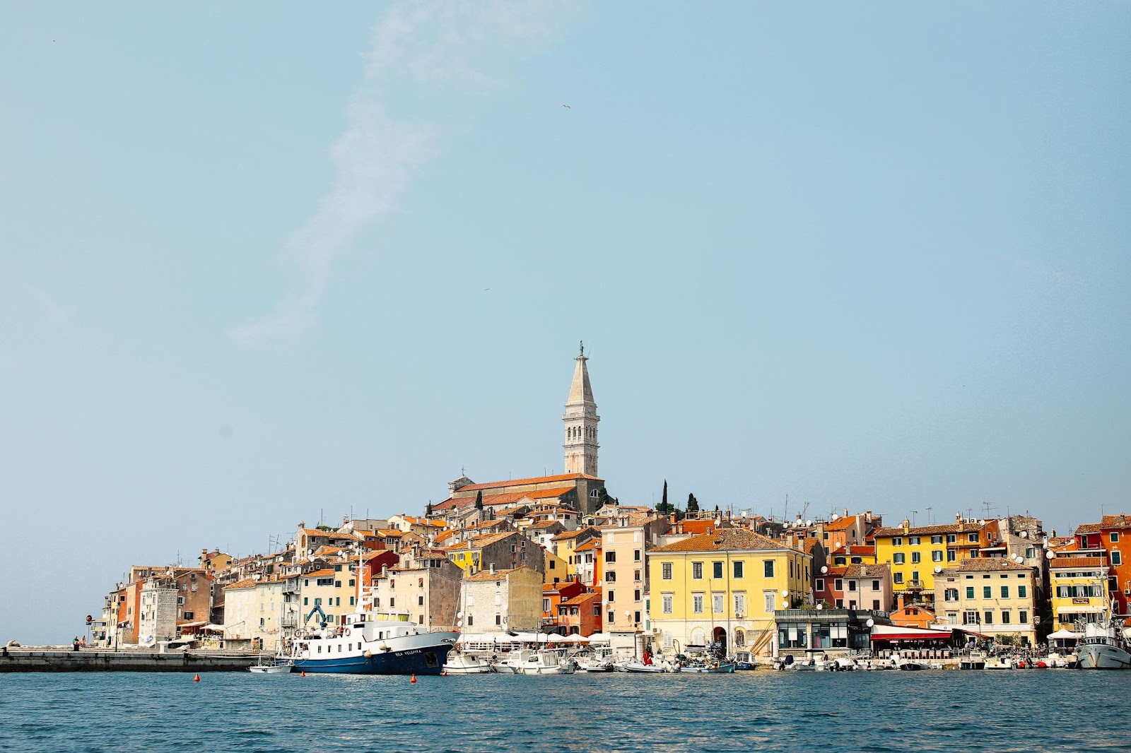 Colorful coastal town with a church tower, boats along the waterfront, and a clear blue sky.