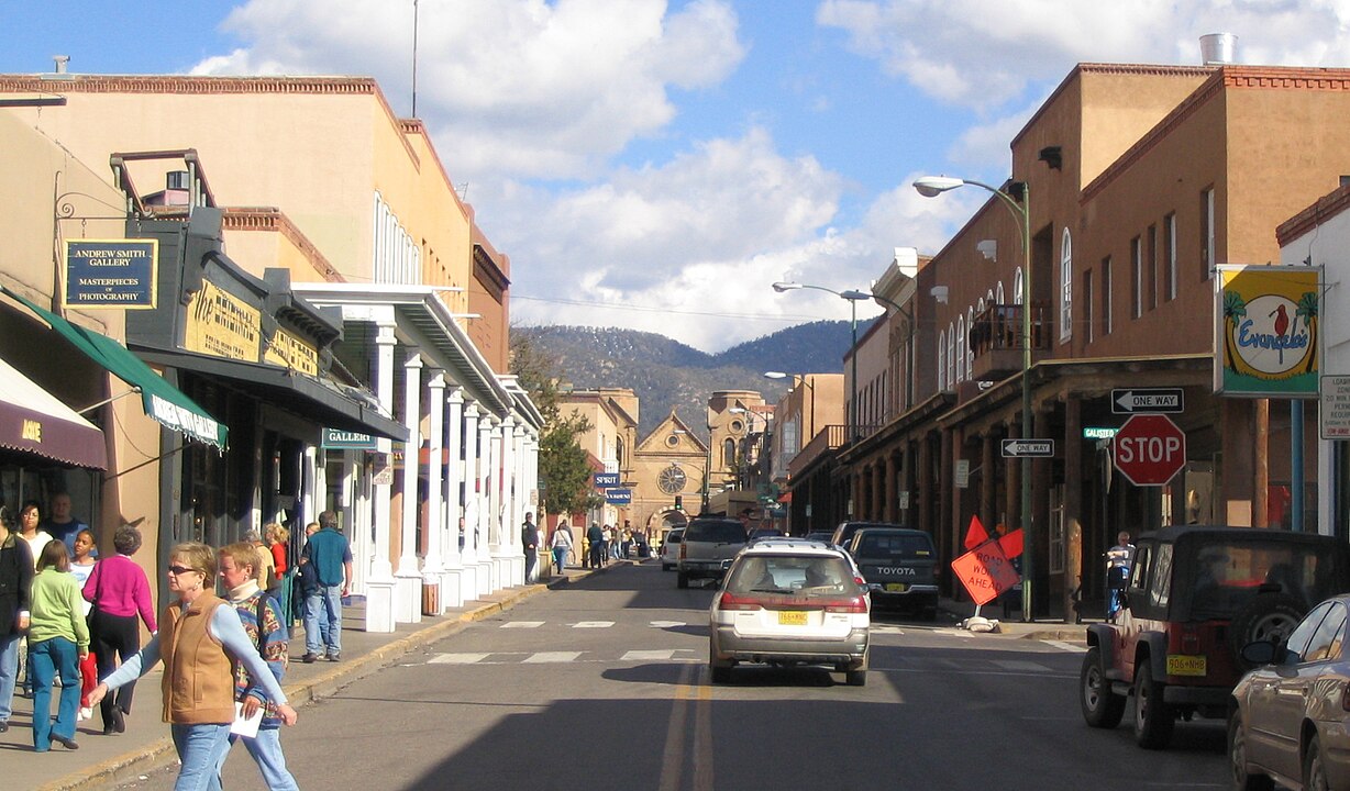 Downtown street with cars, pedestrians, shops, and mountains under a partly cloudy sky.