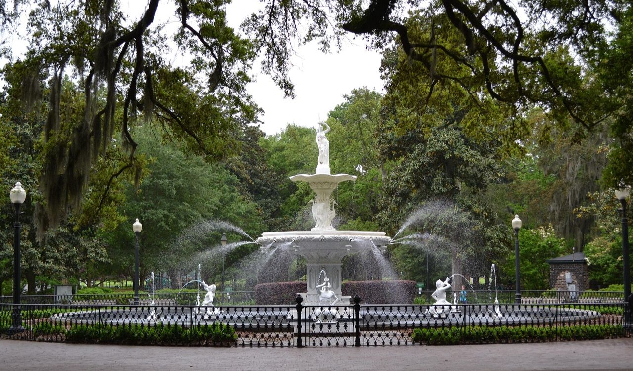 A large white multi-tiered fountain with statues sprays water in a tree-lined park enclosed by a black metal fence.