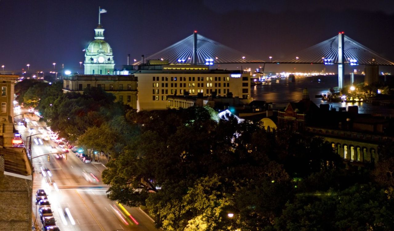 Nighttime city street with car light trails, a domed building, and a lit cable-stayed bridge spanning water.