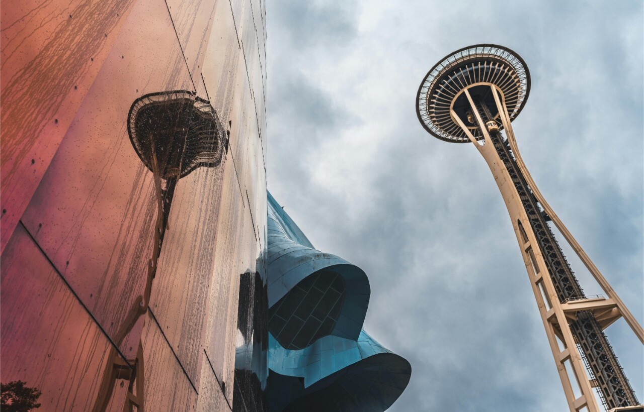 The Space Needle in Seattle is reflected on the shiny metallic surface of a nearby building beneath a cloudy sky.