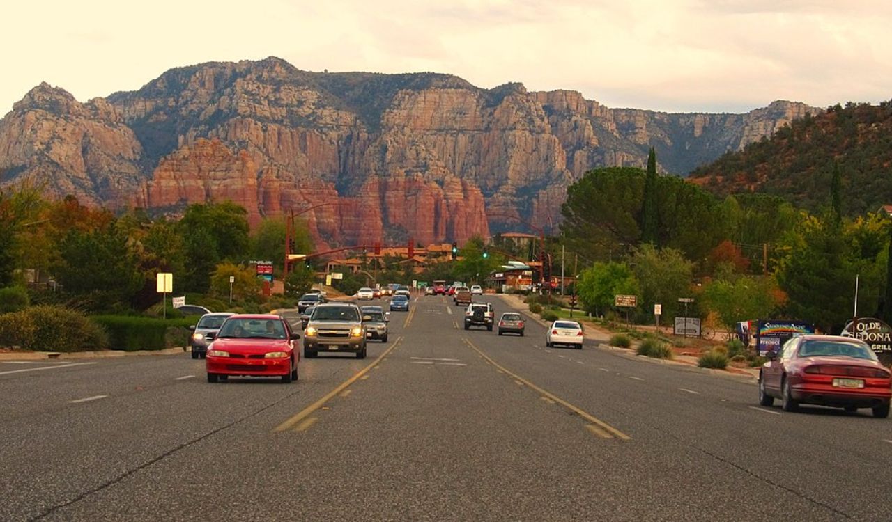 Cars drive along a tree-lined road bordered by small buildings, with large rocky mountains and a partly cloudy sky in the background.