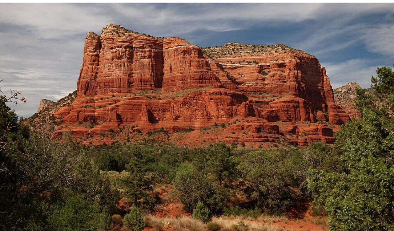 A large, layered red rock mesa rises above green shrubs and small trees beneath a partly cloudy sky.
