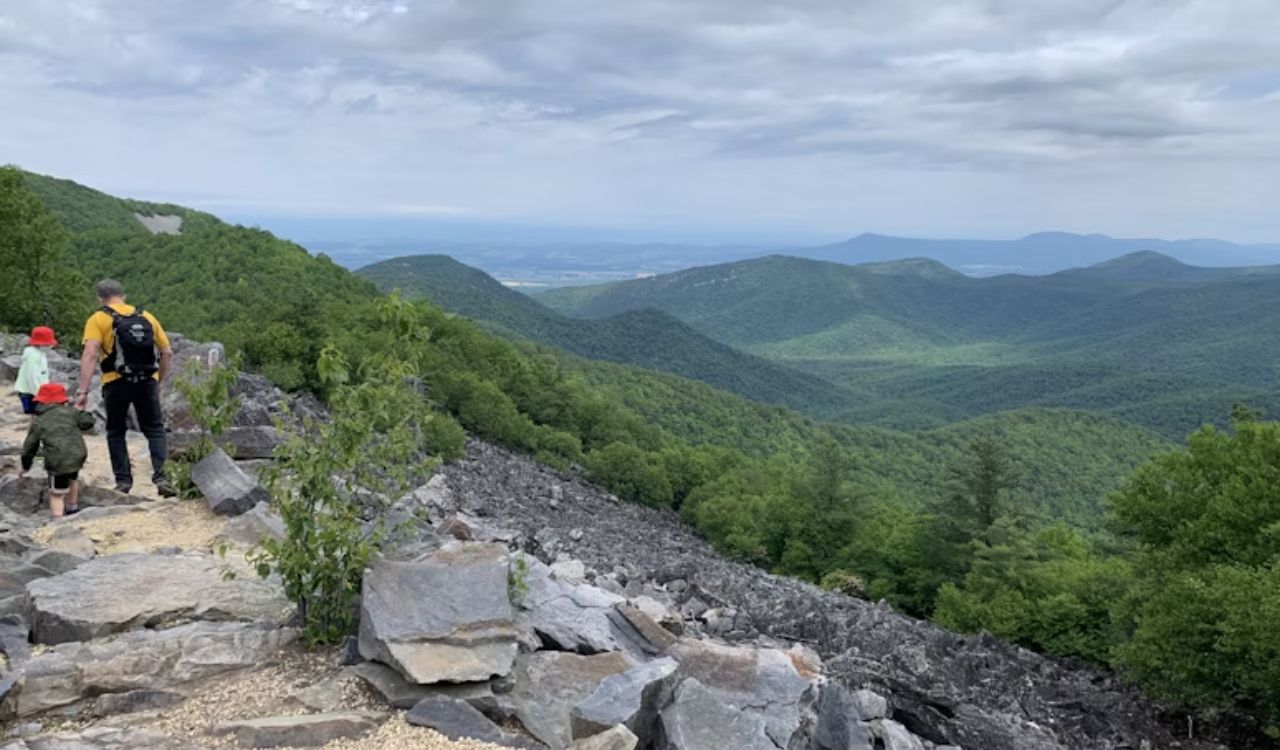 Three people hike a rocky mountain trail amid green hills and distant mountains beneath a cloudy sky.