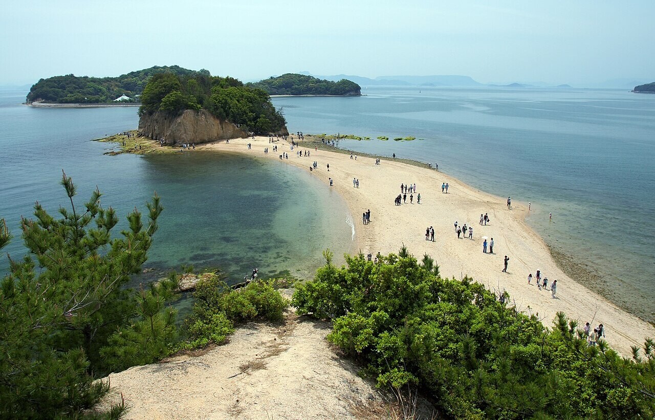 A sandbar links two green islands in calm blue water, with people walking along it under a clear sky.