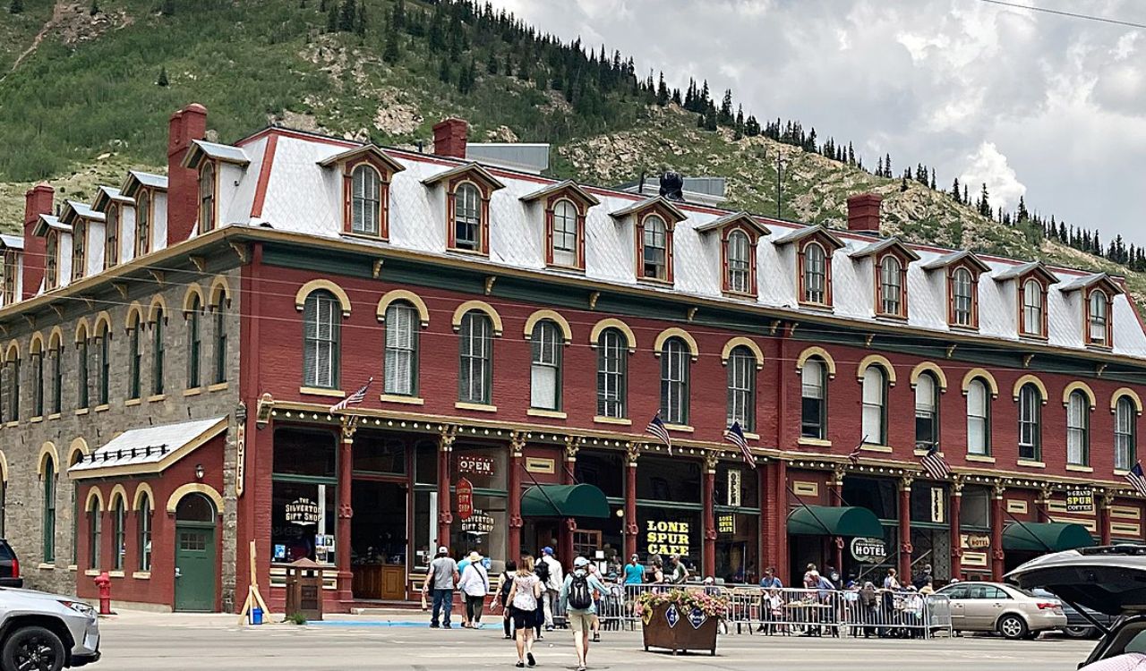 Historic brick building with dormer windows, ground-floor shops, outdoor café seating, and pedestrians, set against a cloudy sky and forested hillside.