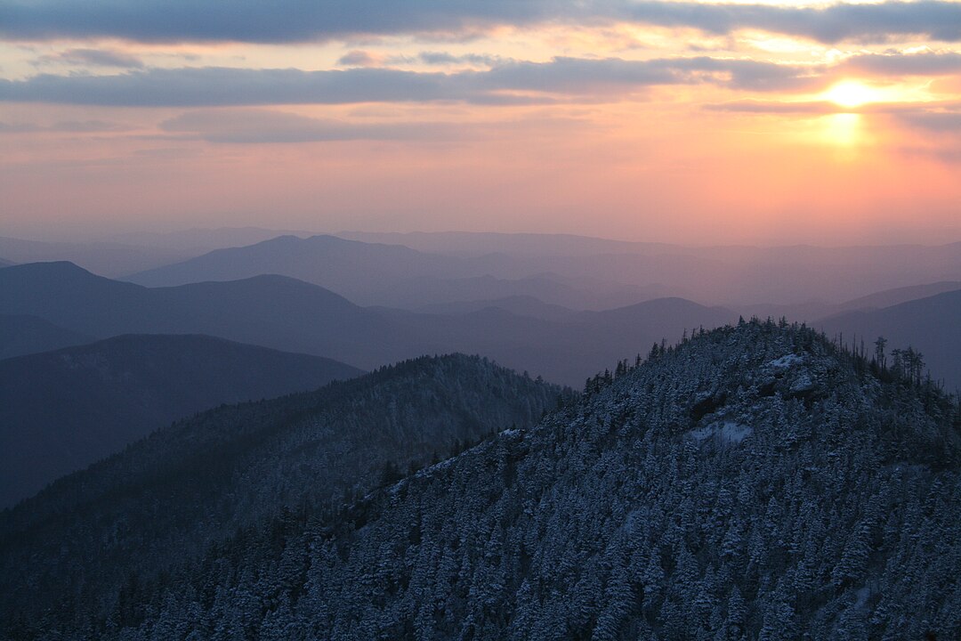 Snow-covered mountain ridges are softly illuminated at sunset beneath a cloudy sky with the partially visible sun.