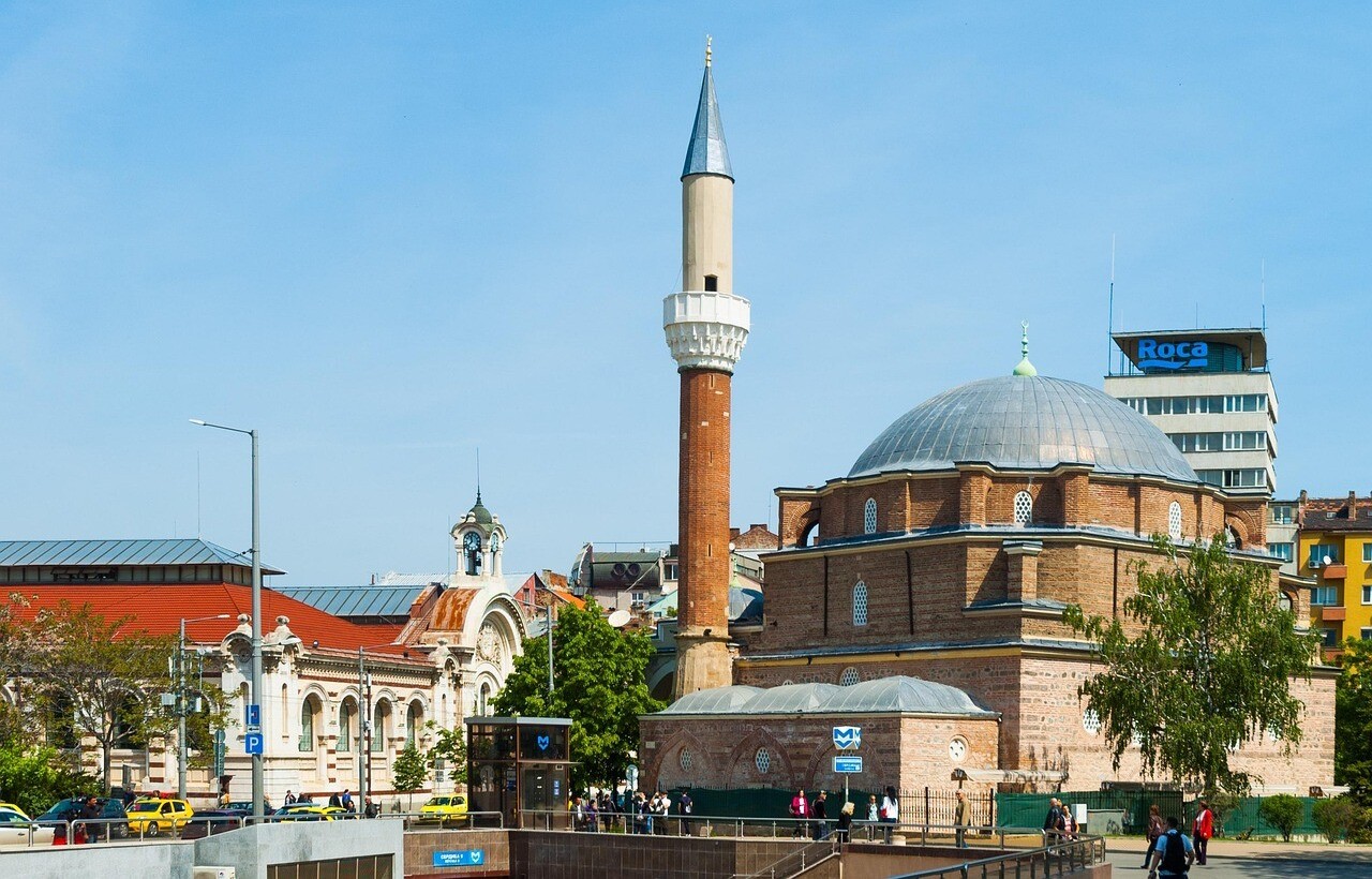 A historic mosque with a tall minaret stands amid urban buildings, trees, and pedestrians on a sunny day.