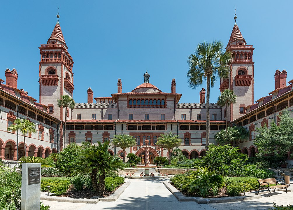 A historic red-brick building with towers and arches stands amid palm trees and gardens beneath a clear blue sky.