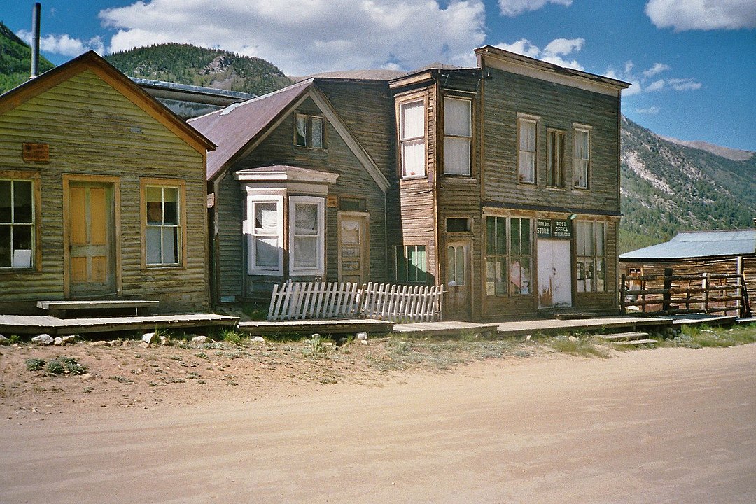 Weathered old wooden buildings line a dirt road in a rural mountain area under partly cloudy skies.