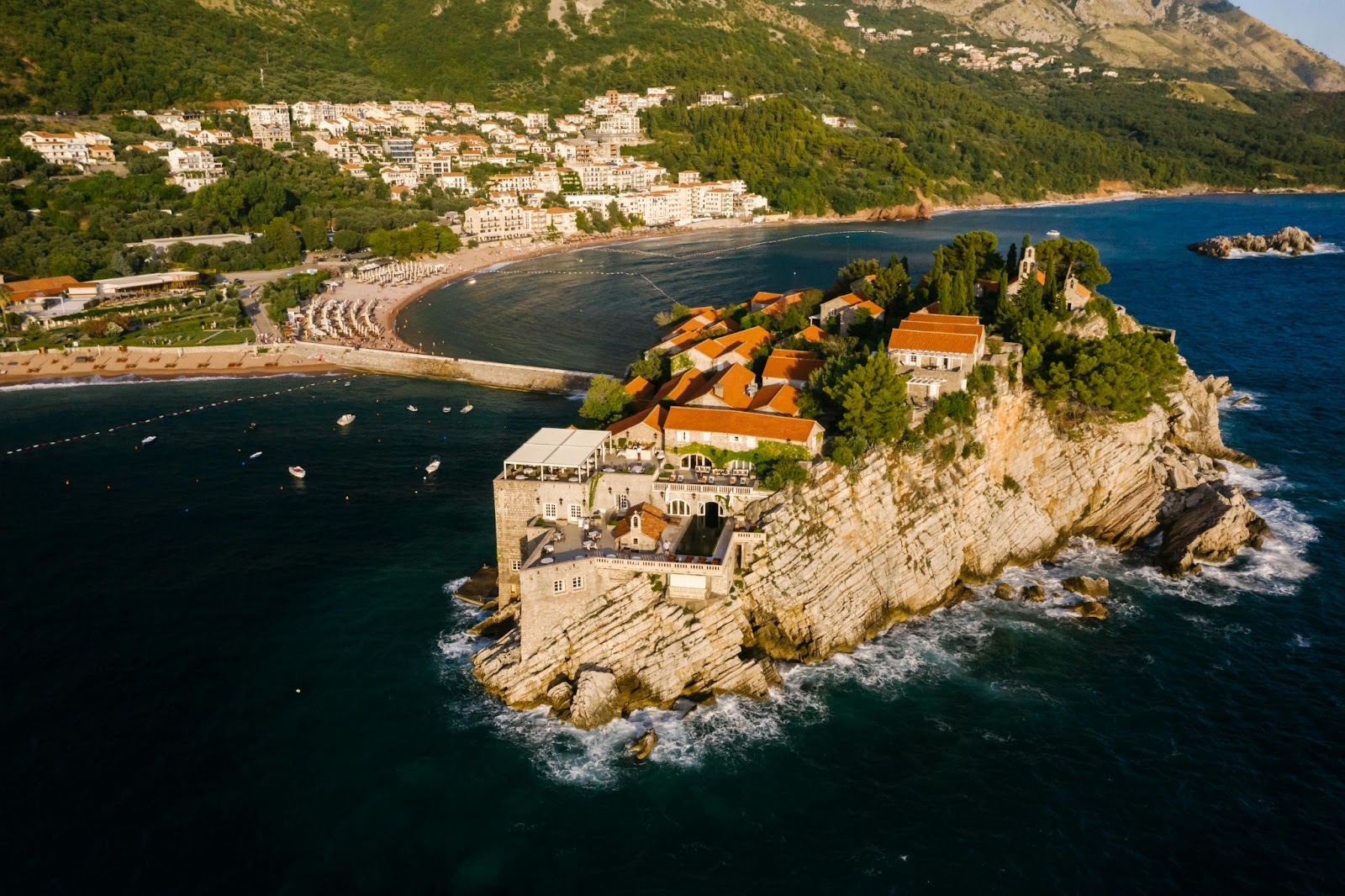 A small island with red-roofed buildings, surrounded by sea and linked to a coastal town by a narrow land strip, is seen from above.