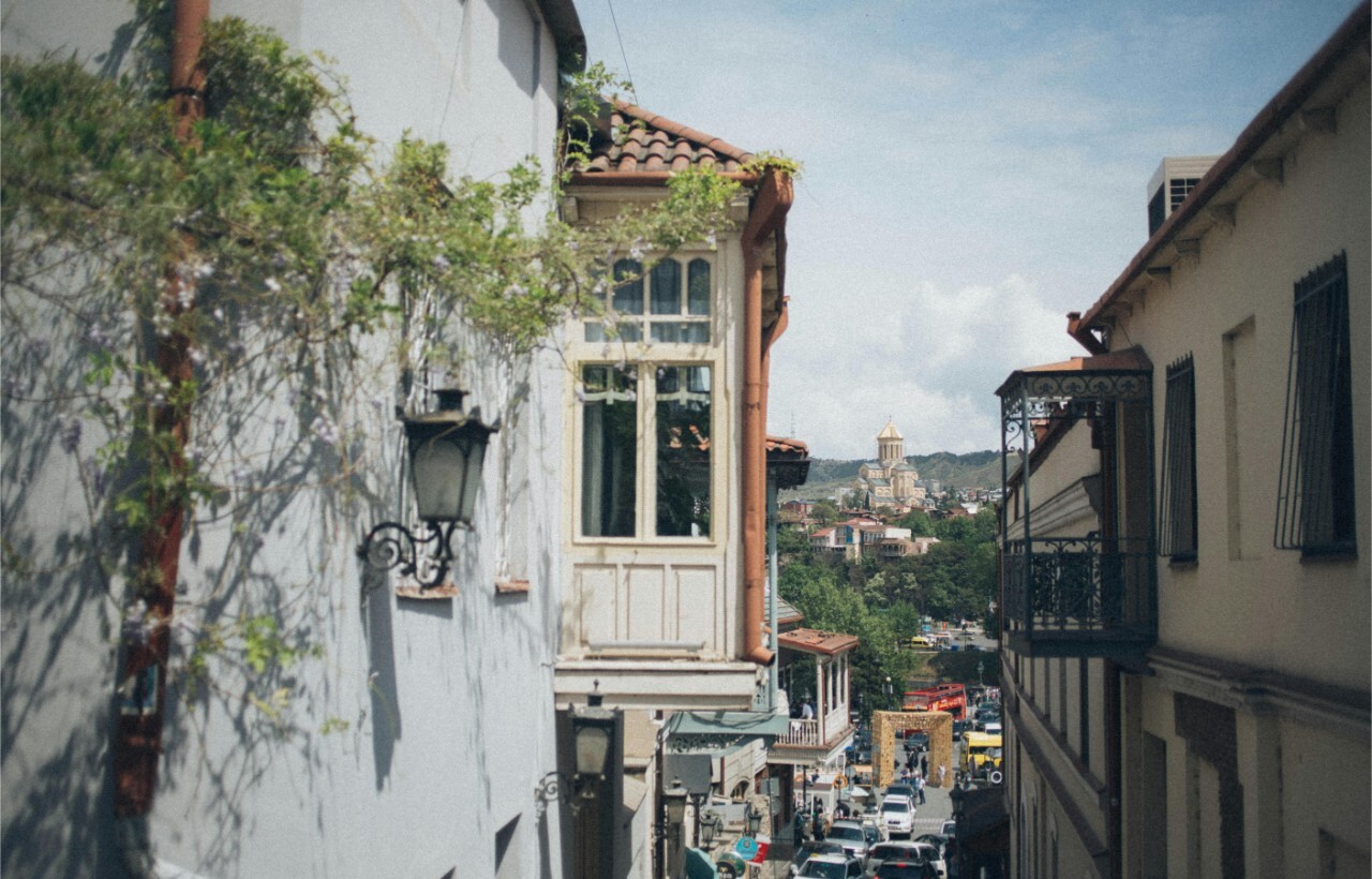 Narrow street lined with buildings, climbing plants, a hanging lantern, and a distant church dome beneath partly cloudy skies.