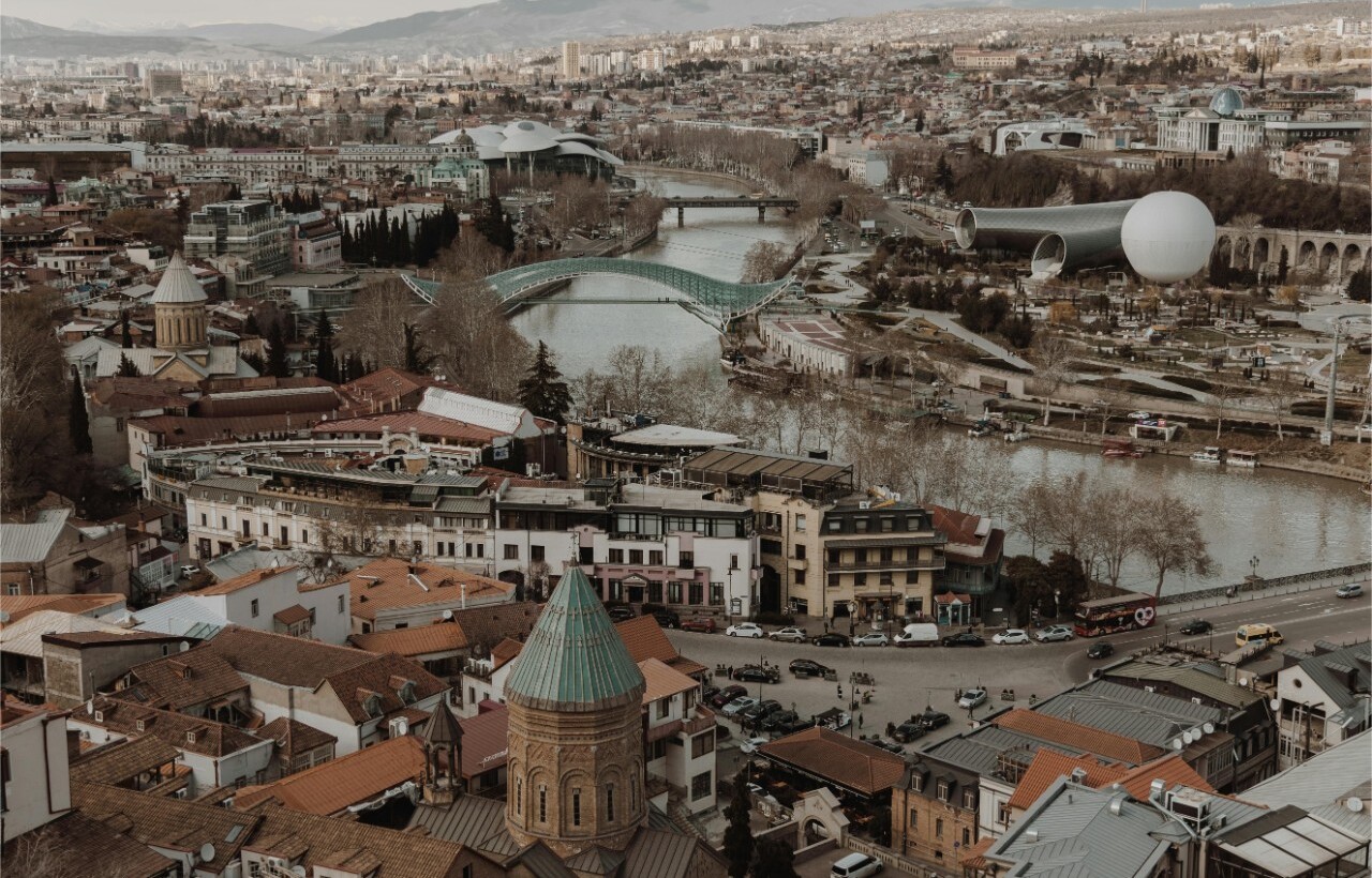 Aerial view of a city featuring a river, modern bridges, historic buildings, and distant mountains beneath an overcast sky.