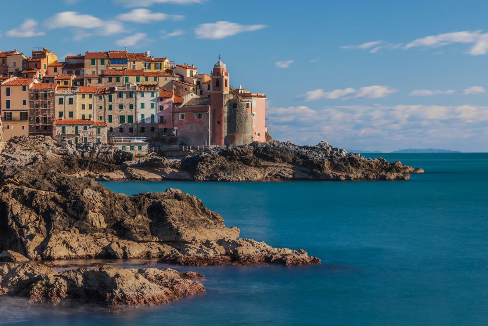 A coastal village with colorful buildings and a church perches on rocky cliffs above a calm blue sea beneath a partly cloudy sky.