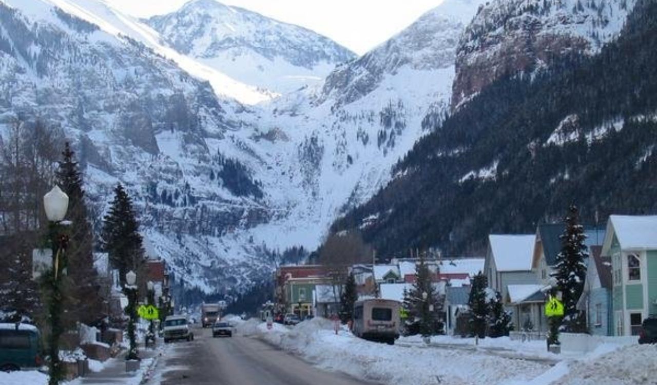 A snow-covered street winds through a small town with houses and trees, set against tall, snowy mountains.