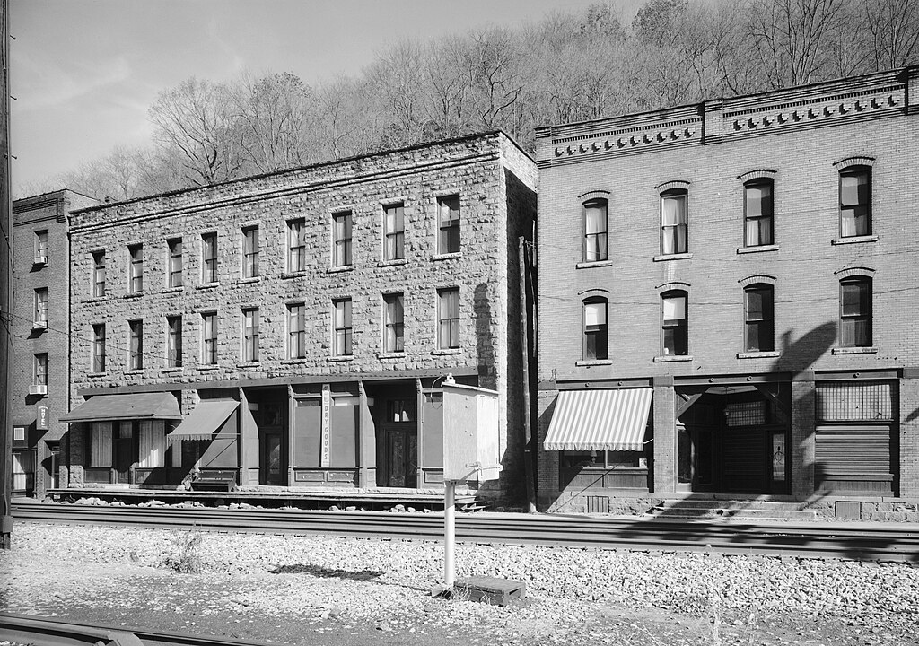 Black and white photo of two three-story brick buildings with covered storefronts behind railroad tracks, with a wooded hill in the background.
