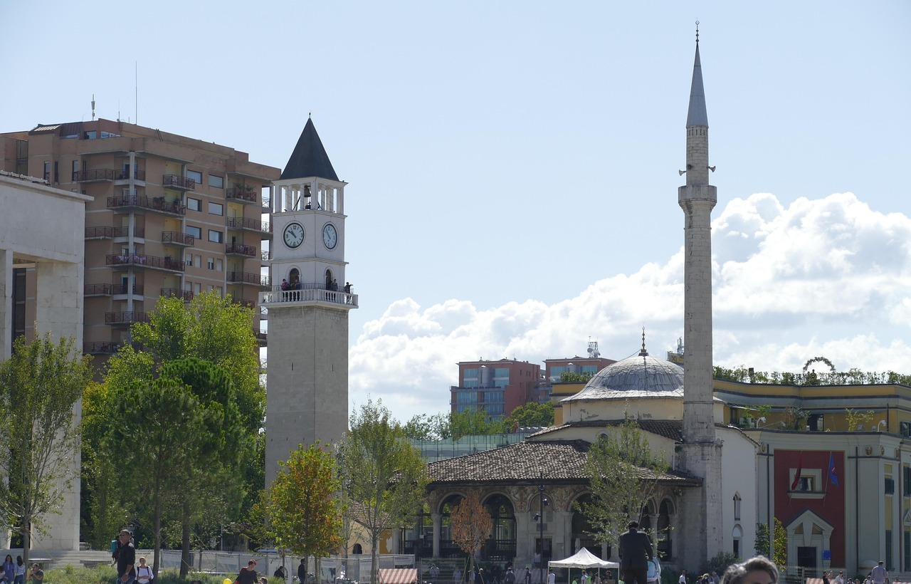 Cityscape with a clock tower, mosque and minaret, residential buildings, and greenery under a partly cloudy sky.