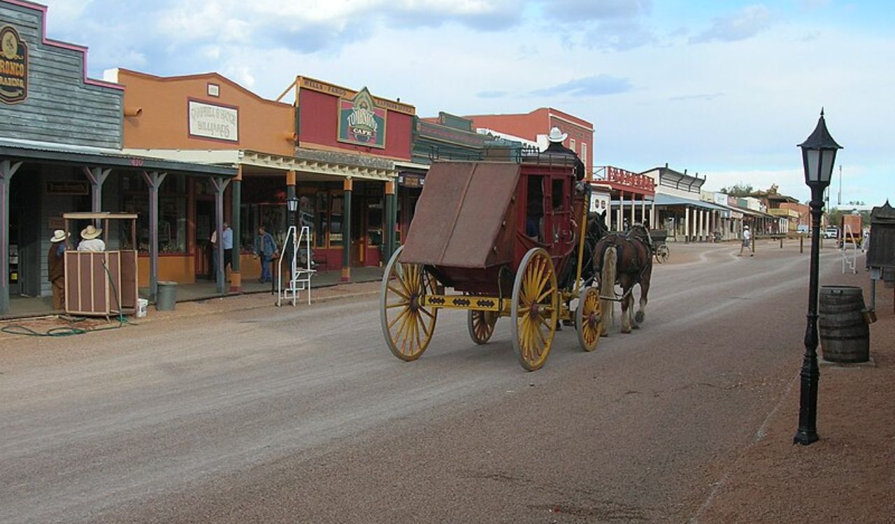 A horse-drawn stagecoach travels down a wide dirt street lined with old western-style wooden buildings under a partly cloudy sky.
