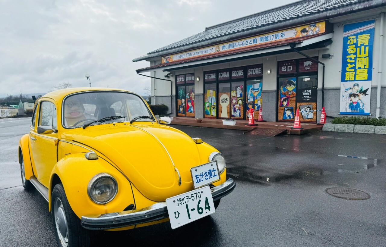 A yellow vintage Volkswagen Beetle is parked before a building adorned with anime-themed banners on a cloudy day.