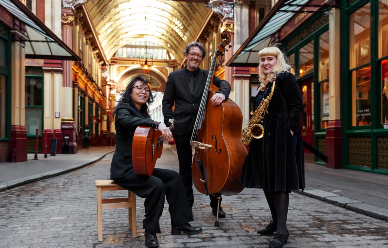 Three musicians pose in an ornate covered market with a guitar, double bass, and saxophone.