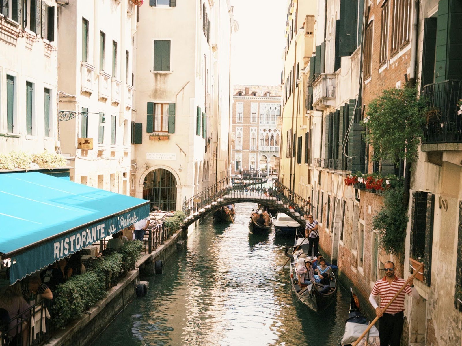 A narrow Venetian canal with gondolas and people passes under a blue "RISTORANTE" awning on the left, flanked by tall buildings and a small arched bridge in the background.