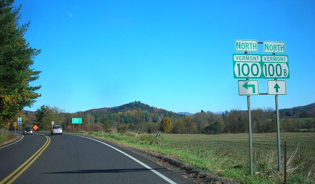 Two Vermont highway signs indicate Route 100 North (left) and Route 100B North (straight) on a rural road with cars, trees, hills, and a clear blue sky.