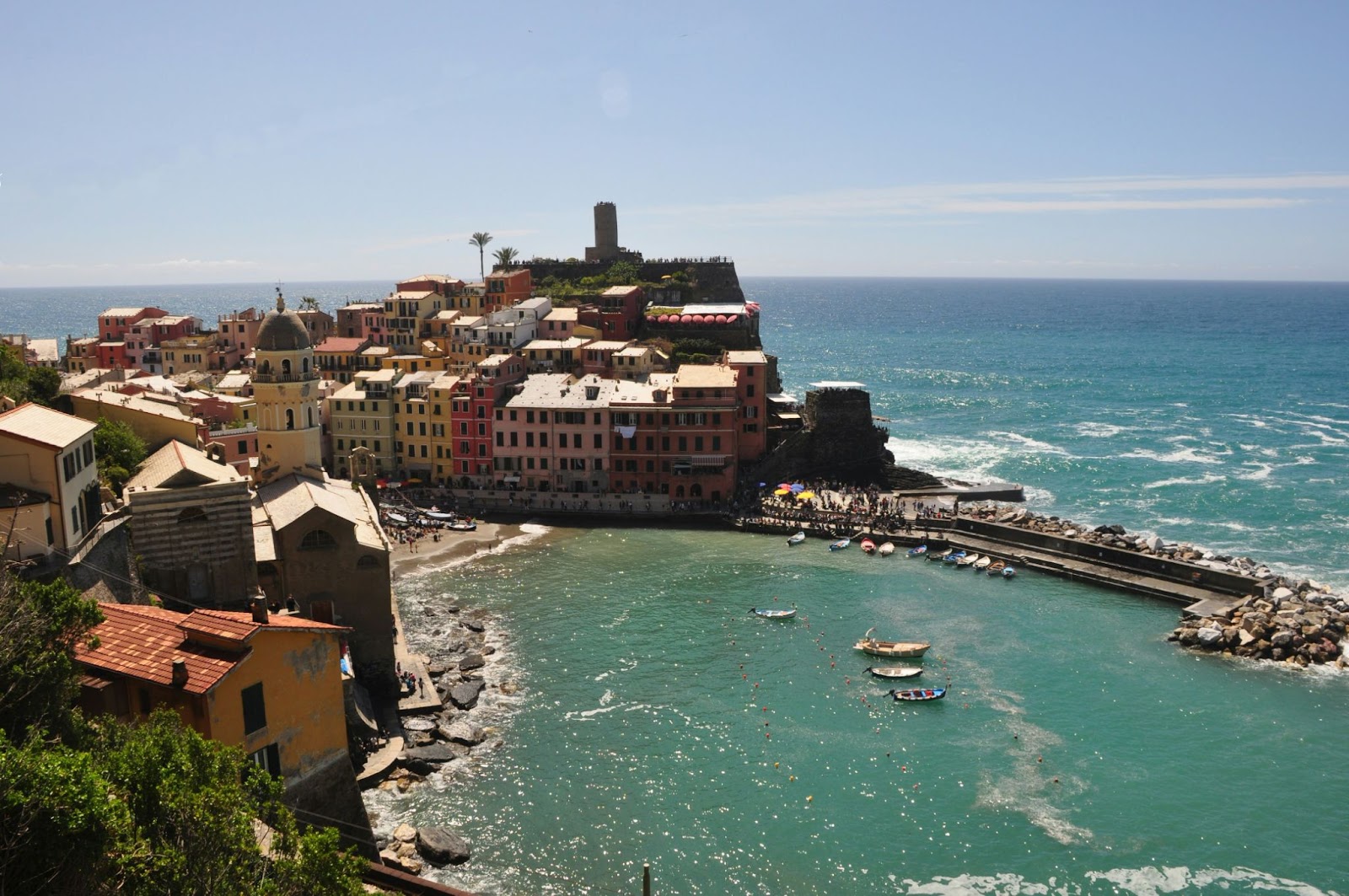 Colorful seaside buildings line a curved harbor of small boats beside the blue sea under a clear sky.